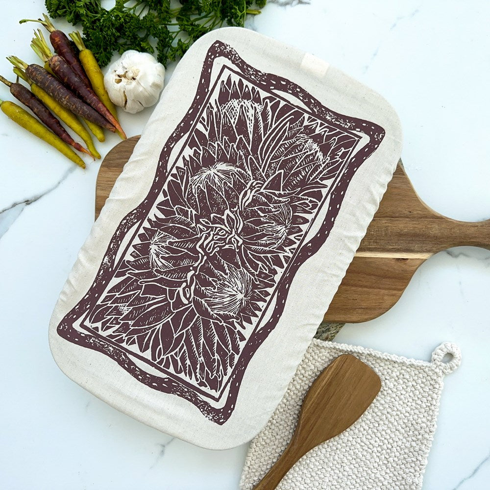 A cloth casserole dish cover with a Protea floral print design, displayed on a wooden cutting board with vegetables in the background.